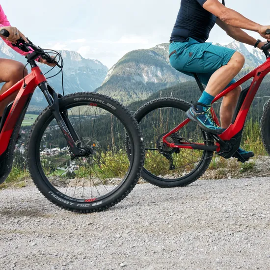 Paar fährt rote E-Mountainbikes auf Schotterweg mit alpiner Bergkulisse