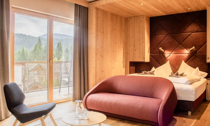 Modern hotel room with wooden interior, red sofa, bed and balcony view of forested mountains