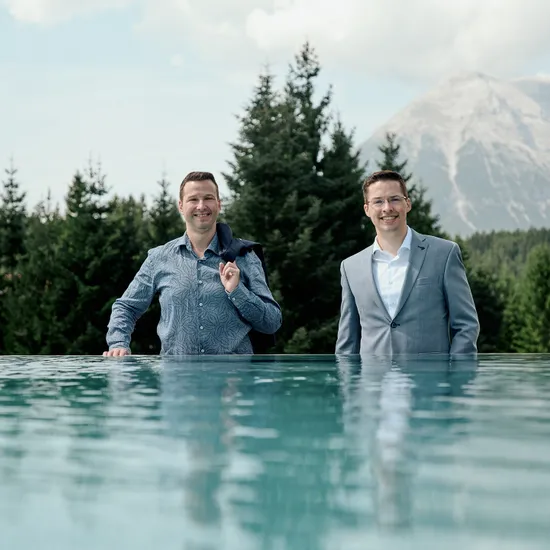 Two smiling businessmen by an infinity pool with forest and mountain backdrop