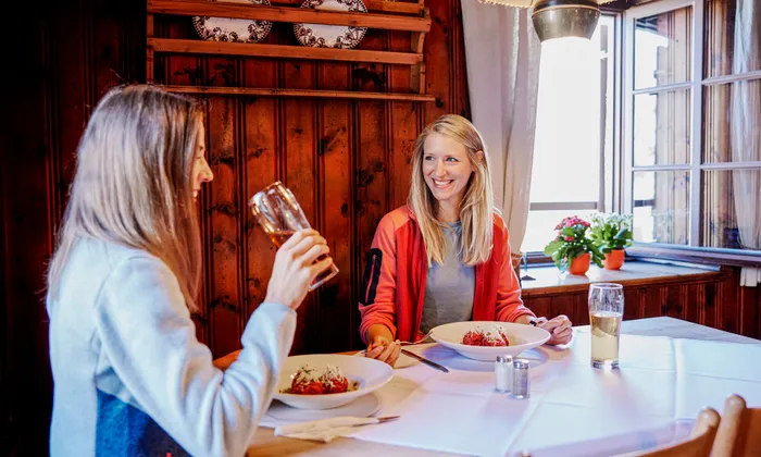 Two women enjoying pasta and drinks in a cozy wooden restaurant with natural light