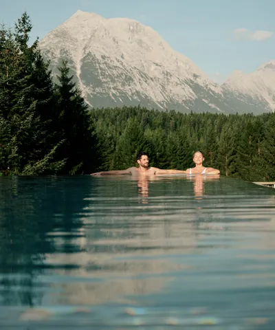 Paar entspannt im Infinity-Pool mit Blick auf Berge und Wald im Sommer