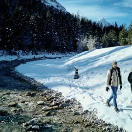 Zwei Personen gehen an einem verschneiten Flussufer im Winterwald mit Bergblick entlang