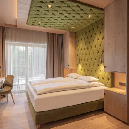Modern hotel room with green upholstered bed and wood accents, natural light from balcony door