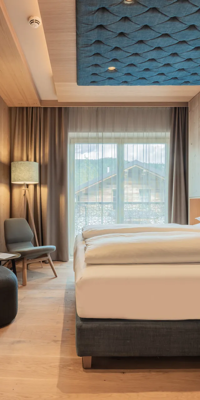 Modernes Hotelzimmer mit Holzelementen, großem Bett, blauer Polsterwand, Schreibtisch und Bergblick