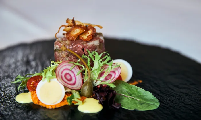 Gourmet beef tartare with quail eggs, beet slices, greens and sauce on black slate plate