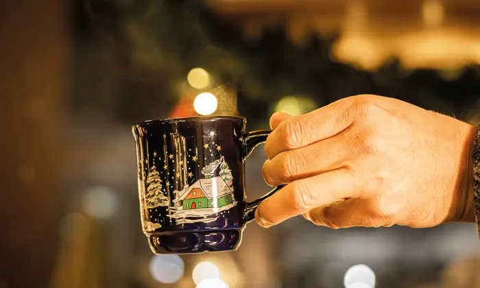 Hand holding steaming Christmas mug with snowy cabin design at festive market