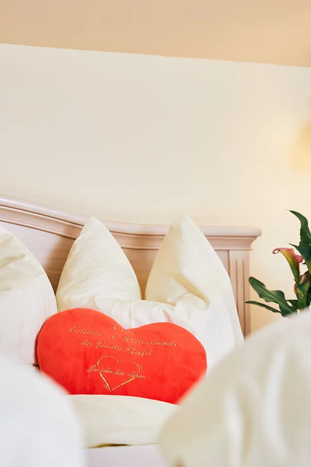 Gemütliches Hotelbett mit herzförmigem rotem Kissen und frischen Calla-Lilien neben Wandlampe